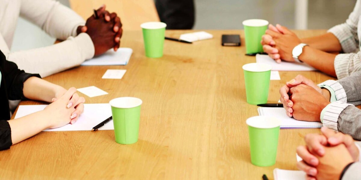 Close-up of people seated around a meeting table with folded hands, notebooks, pens, and green paper cups.